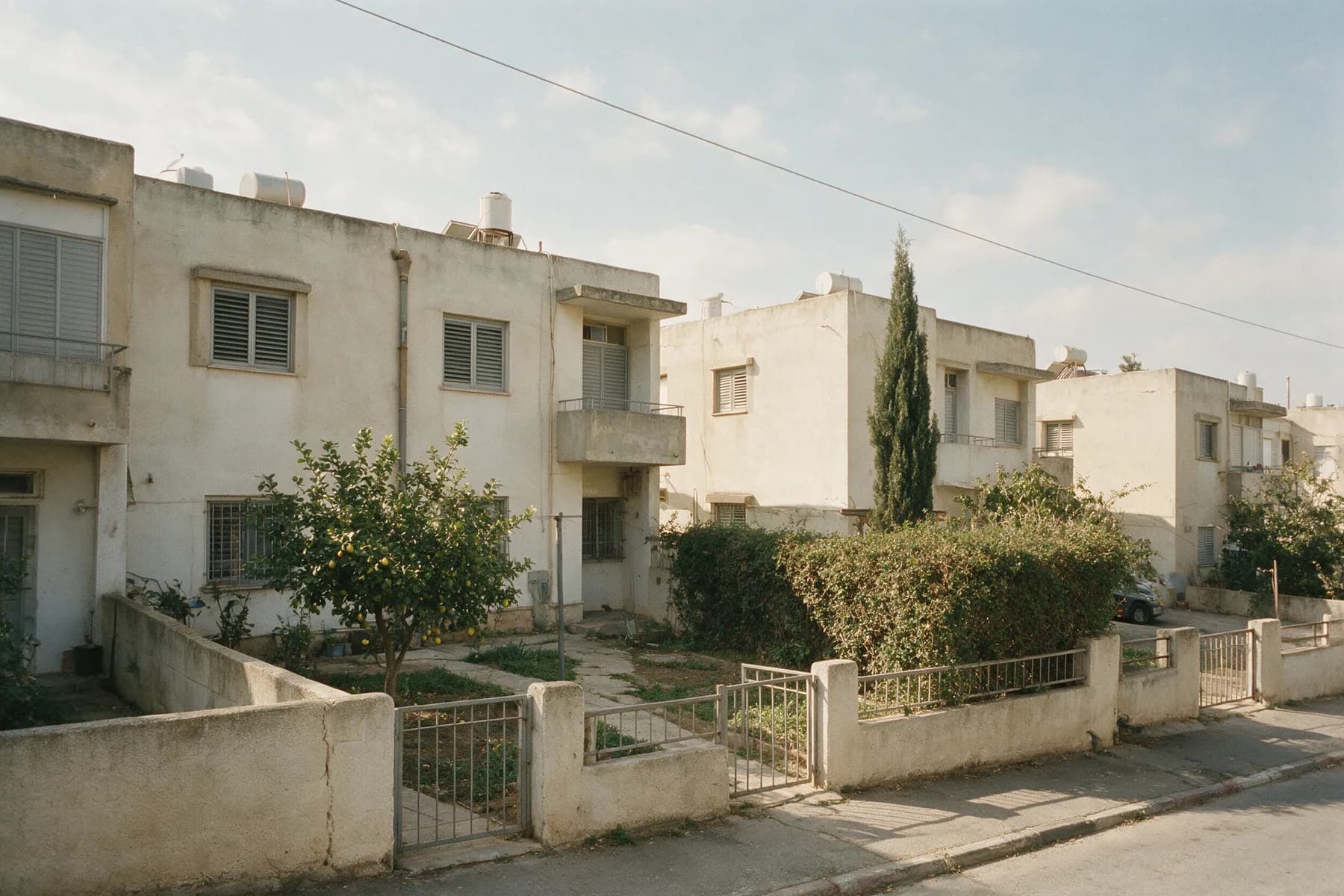 רחוב של בתים נמוכים מטויחים בשתי קומות, גגות שטוחים עם דודי שמש, תריסי אלומיניום, גדרות חצר נמוכות עם שערי מתכת ועץ לימון בחצר